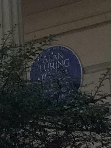 Close-up of Alan Turing’s blue plaque on Warrington Crescent, partially obscured by tree branches.